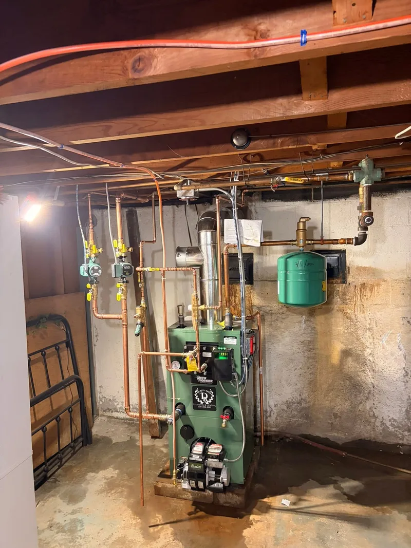 A green oil-fired boiler installed in a basement, with its burner at floor level, a flue rising to the ceiling, copper near-boiler piping with zone circulators, and a green expansion tank hung on the wall beside it.
