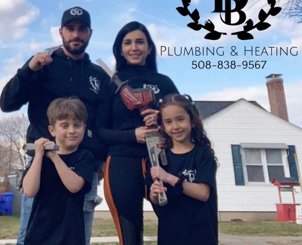 Two adults and two children in matching De Rosa Bros work clothing standing on a front lawn, each holding a pipe wrench, with a clapboard house and a bare tree behind them. The De Rosa Bros wreath logo, the words Plumbing and Heating, and a phone number are set into the top right of the picture.