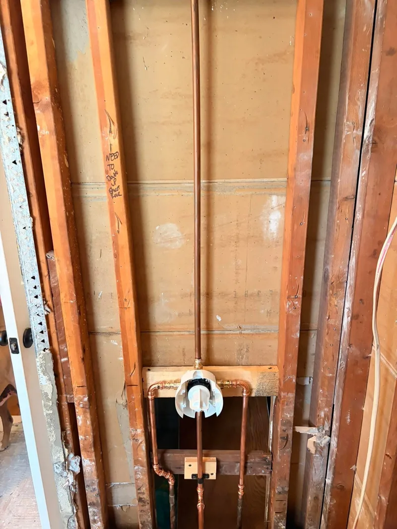 A shower mixing valve and its copper riser roughed into a stud wall in an older bathroom, with the plaster stripped back to the framing either side.
