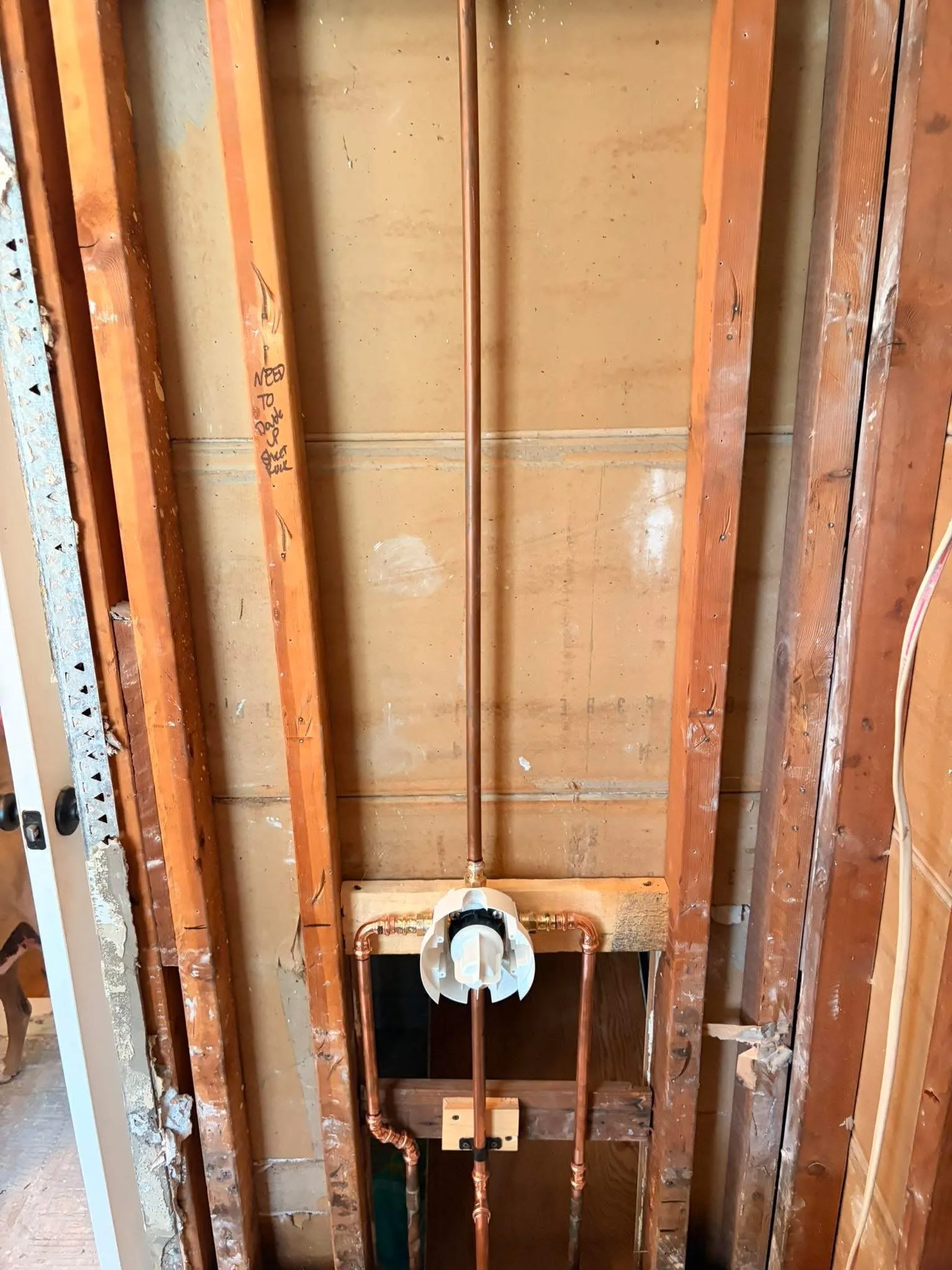 A shower mixing valve and its copper riser roughed into a stud wall in an older bathroom, with the plaster stripped back to the framing either side.