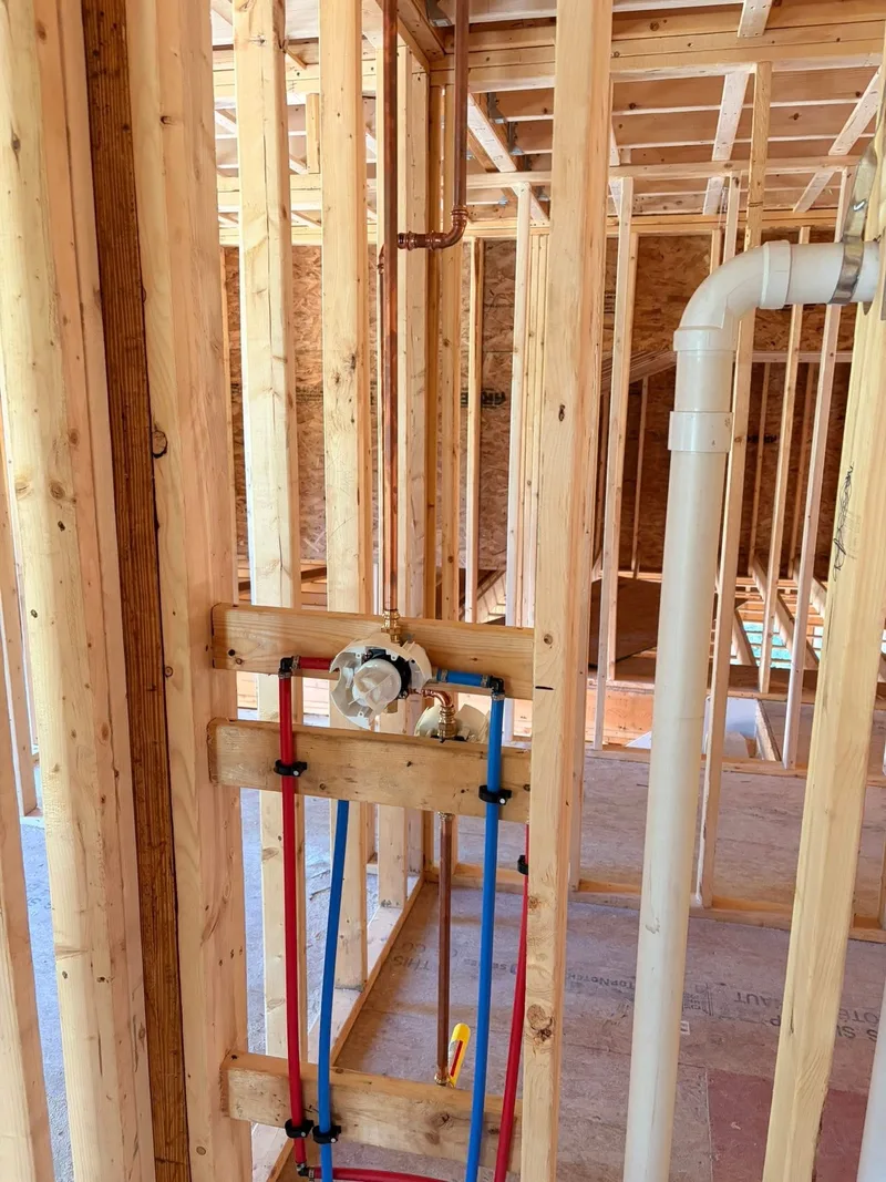 A shower mixing valve mounted on a timber brace between studs in a house under construction, with red and blue plastic supply lines and a copper riser run up to it and a white plastic waste stack alongside.