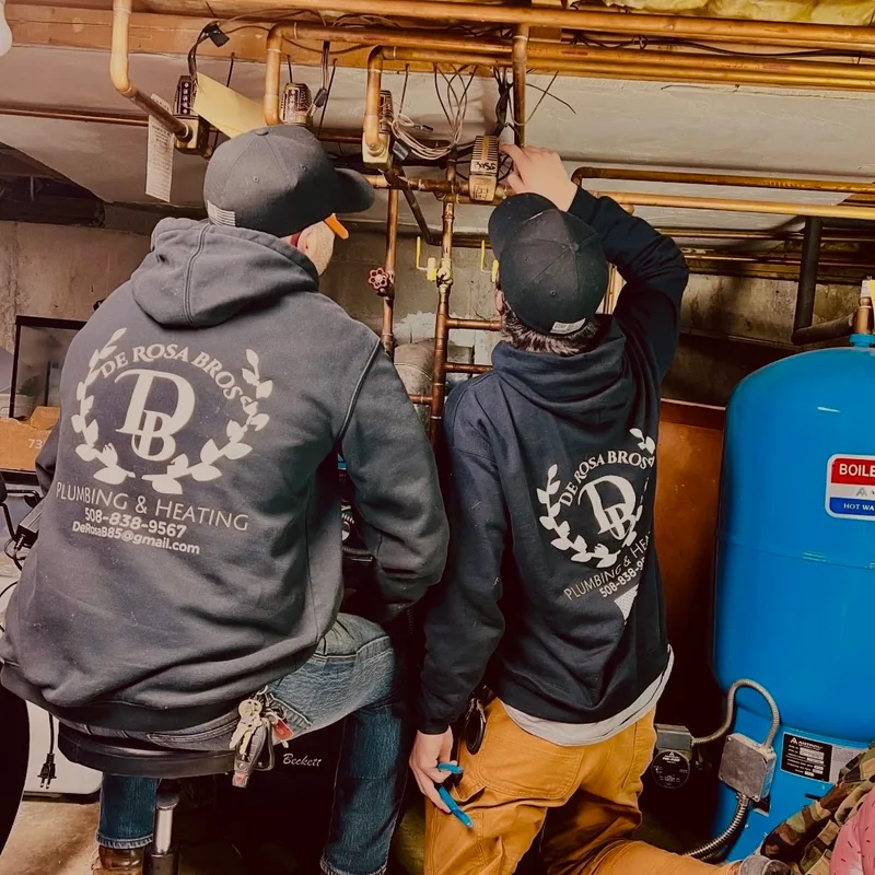 Two plumbers in branded hoodies, seen from behind, working on overhead copper pipework in a basement beside a blue expansion tank.
