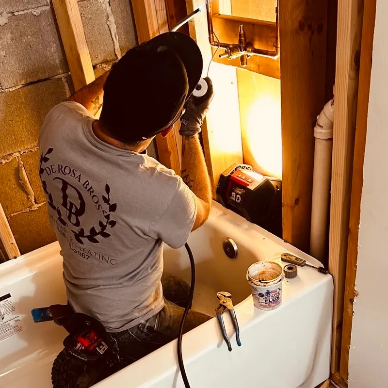A plumber in a De Rosa Bros shirt working inside an opened wall to connect a new bathtub.