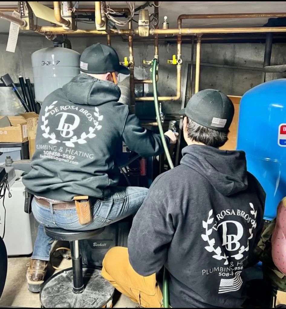 Two plumbers in De Rosa Bros hoodies working on a basement heating system beside a blue expansion tank.