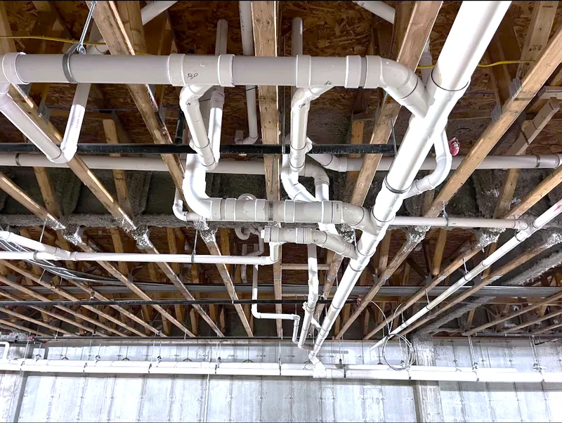 A waste stack and branch lines roughed in overhead between floor joists.