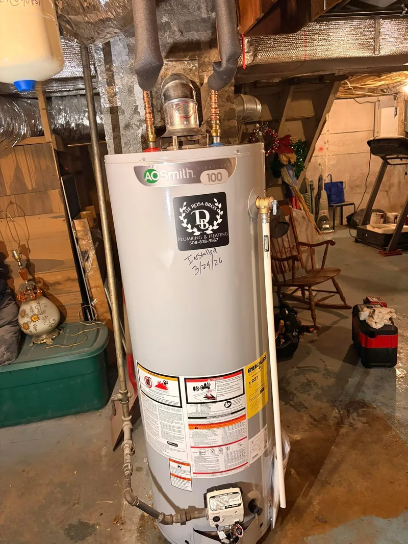 A newly installed gas water heater standing in a basement with its flue elbow, copper supply connections and temperature and pressure relief pipe fitted, and a De Rosa Bros sticker on the front of the tank.