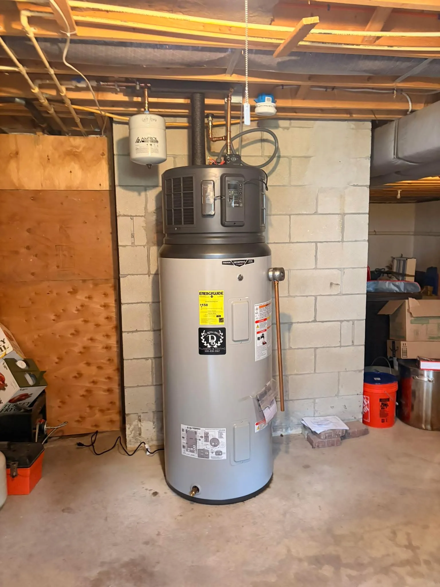 A heat-pump water heater newly installed against a painted block basement wall, with its compressor unit on top of the tank, a small expansion tank on the pipework above and a De Rosa Bros sticker on the front.