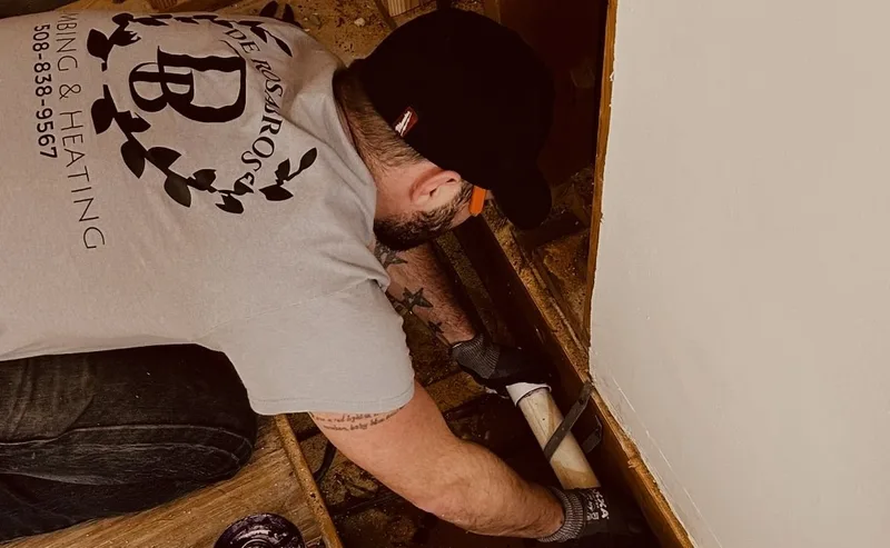 A plumber in a De Rosa Bros shirt reaching into an opened floor to work on pipework below.
